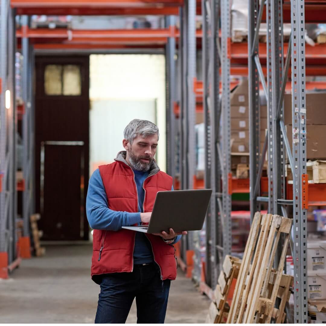 Man in a warehouse using a laptop, wearing a red vest.
